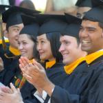A group of graduating master's students at their commencement ceremony, wearing black caps and gowns with gold master's hoods.
